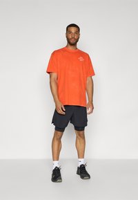 Orange bomulds T-shirt med rund halsudskæring og trykt tekst, parret med sorte træningsshorts og sorte sneakers.