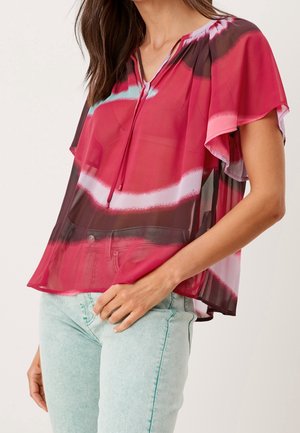 Žena nosí volnou krátký rukáv červenou a růžovou vzorovanou halenku s zavazováním u výstřihu a světle modré džíny.