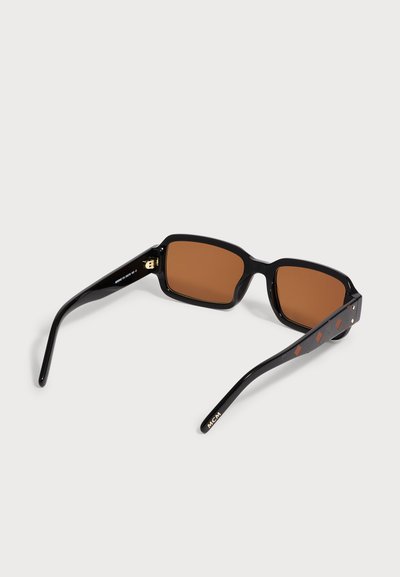 Lunettes de soleil rectangulaires noires avec des verres marron, des branches décorées et un logo doré sur la branche reposant sur une surface blanche.