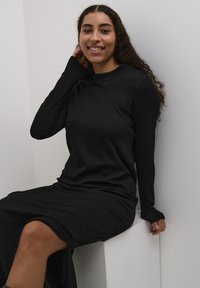 Black long-sleeve dress made from textured fabric, featuring a crew neck and a high slit on one side, sitting on a white pedestal.