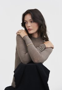 Woman with dark wavy hair wearing a brown long-sleeve top, gold rings, and bracelet, arms crossed resting on black fabric.