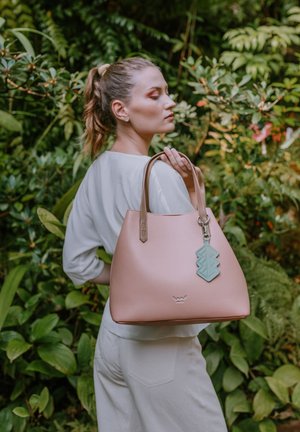 Frau mit Pferdeschwanz, die ein weißes Outfit trägt und eine große rosa Handtasche mit einem blauen geometrischen Schlüsselanhänger in einem üppigen grünen Garten hält.