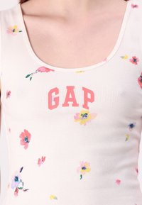 Débardeur blanc à côtes avec des imprimés floraux en rose, jaune et bleu, arborant un logo "GAP" rose à l'avant.