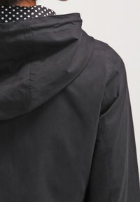 Czarna kurtka z kapturem o matowej teksturze. Zawiera widok z tyłu, podkreślający bezszwowy design i lekko zakrzywiony dół. Koszula w kropki widoczna pod spodem.