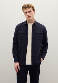 Donkerblauw overhemd met knopen en twee borstzakken, gemaakt van een gladde stof. Gedragen over een lichtgekleurde T-shirt met ronde hals.