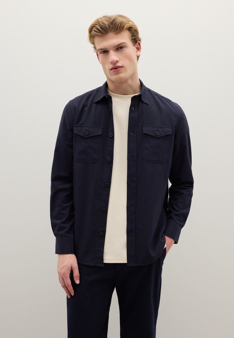 Donkerblauw overhemd met knopen en twee borstzakken, gemaakt van een gladde stof. Gedragen over een lichtgekleurde T-shirt met ronde hals.