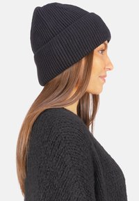 Bonnet tricoté noir à côtes avec un revers, présentant une surface texturée, un tissu doux et un ajustement serré, porté de biais.
