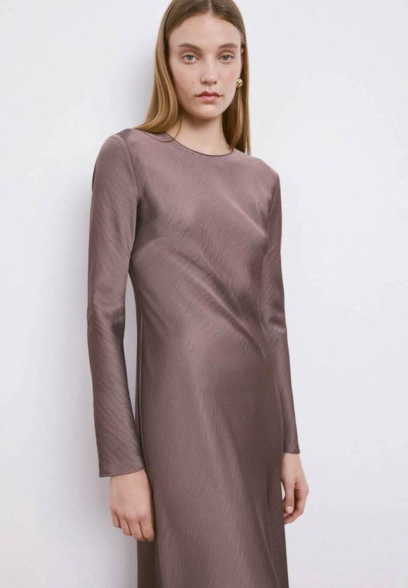 Femme aux cheveux blonds raides portant une robe en satin mauve à manches longues, debout devant un fond clair uni.