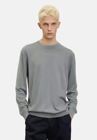 Maglione grigio chiaro a maniche lunghe con scollo rotondo, polsini a costine e una texture liscia, abbinato a pantaloni scuri.
