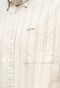 Μπεζ πουκάμισο με κουμπιά, με κάθετες καφέ ρίγες και τσέπη στο στήθος με κεντημένο καφέ κείμενο.