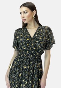 Robe noire florale avec une taille smockée, des manches courtes en tulle et des accents dorés. Présente un décolleté en V et des motifs complexes verts et dorés.