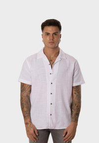 Camisa de manga corta de lino blanco con cuello, que presenta botones marrones en el frente. Tejido texturizado, ajuste relajado, diseño moderno.