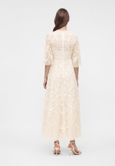 Femme aux longs cheveux bruns portant une robe midi crème brodée de dentelle, manches trois-quarts et sandales à talons beiges, de dos.