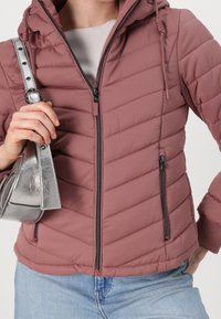 Jasnoróżowa pikowana kurtka z kapturem, z zamkiem błyskawicznym z przodu i bocznymi kieszeniami, ściągaczami przy mankietach oraz gładkim wykończeniem. Srebrna torebka w dłoni.