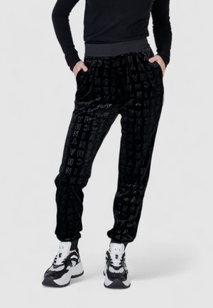 Pantaloni jogger in velluto nero con un motivo a lettere in rilievo, dotati di tasche laterali e una vita aderente, abbinati a sneaker bianche e nere.