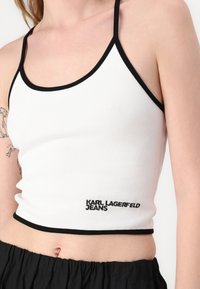Hvit ribbestrikket crop topp med svart kant og justerbare stropper. Har "KARL LAGERFELD JEANS" brodert i svart nær kanten.