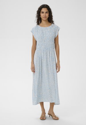 Kobieta o kręconych włosach, ubrana w midi sukienkę w jasnoniebieskie i beżowe wzory z krótkimi rękawami typu kaftan oraz sandałami, stoi na tle jednolitego tła.