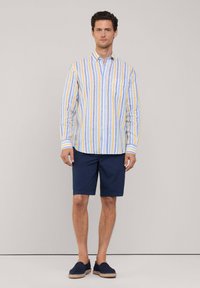 Camisa de botones a rayas en azul claro, amarillo y blanco, combinada con pantalones cortos de color marino. La camisa cuenta con un bolsillo y las mangas enrolladas.