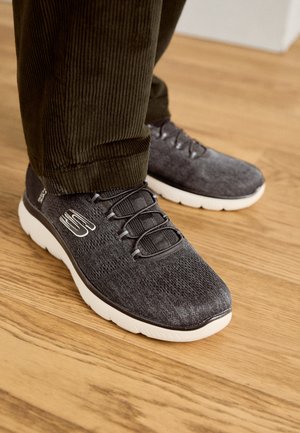 Grå atletik sko med en tekstureret strikoverdel, elastiske snørebånd og en kontrasterende hvid sål, placeret på et trægulv.