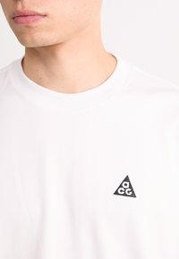 Close-up of a person wearing a white shirt with a small black triangular logo featuring the letters "ACG" on the chest.