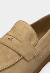 Μπεζ moccasin από σουέτ με ραμμένες λεπτομέρειες και μικρό χρυσό λογότυπο "MK" στο επάνω λουράκι, κοντινή γωνιακή λήψη.