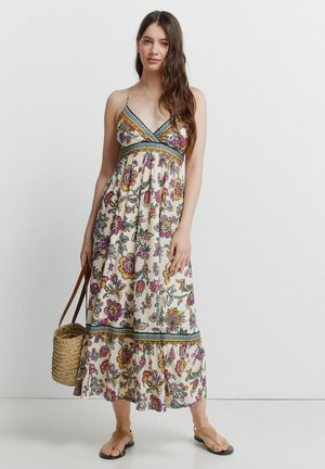 Frau in ärmellosem Blumen-Maxikleid, die eine geflochtene Korbtasche hält und beige flache Sandalen trägt, vor einem einfarbigen Hintergrund.