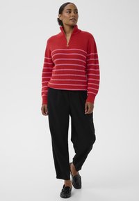 Czerwono-różowy paskowany sweter z prążkowanej dzianiny z kołnierzem na pół-zamek, połączony z czarnymi luźnymi spodniami i czarnymi butami wsuwanymi.