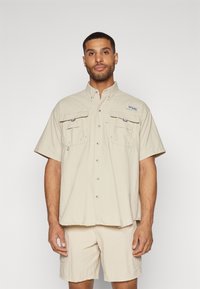 Camicia beige a maniche corte con due tasche sul petto, chiusura con bottoni e una vestibilità leggermente rilassata, abbinata a pantaloni corti coordinati. Materiale leggero.
