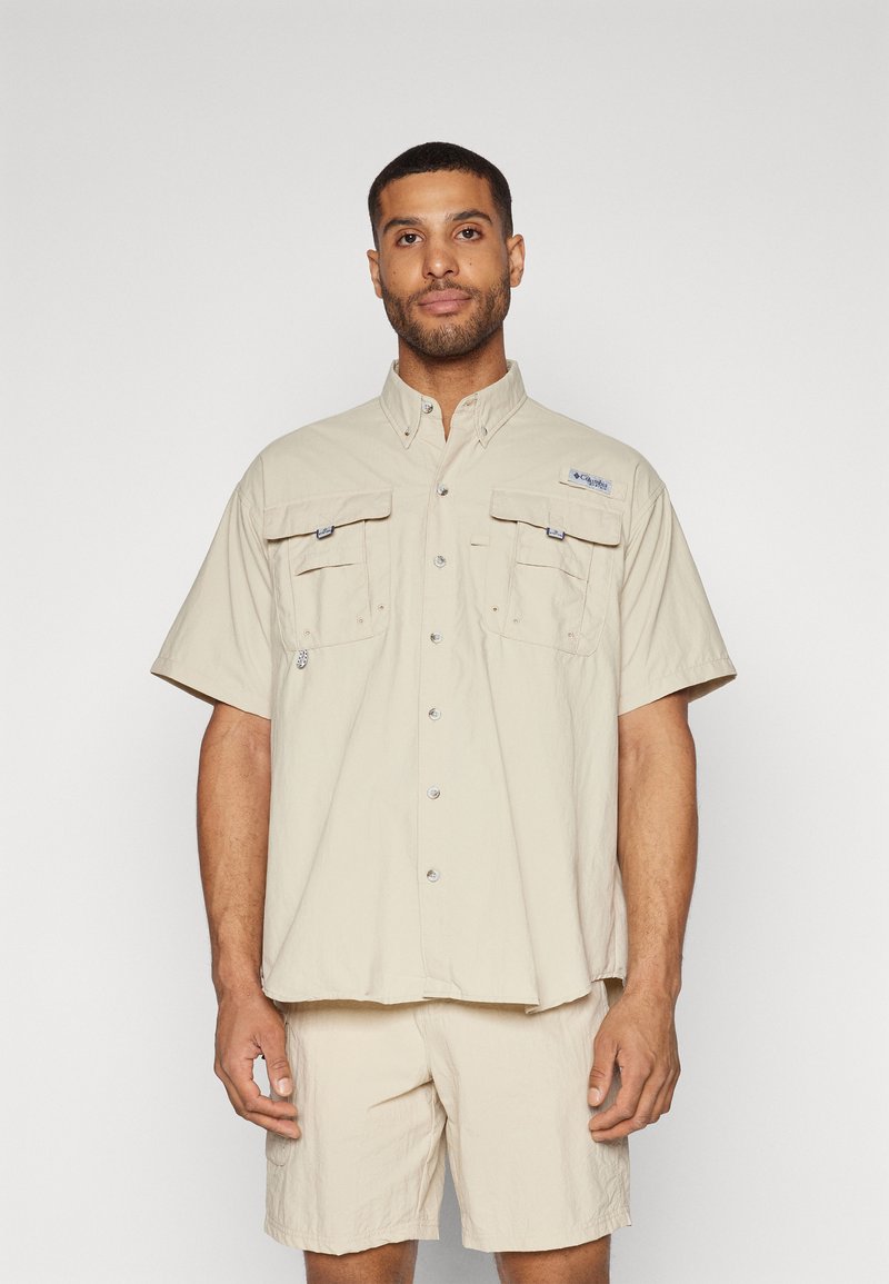 Camicia beige a maniche corte con due tasche sul petto, chiusura con bottoni e una vestibilità leggermente rilassata, abbinata a pantaloni corti coordinati. Materiale leggero.