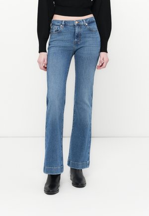 Jean flare bleu en denim, avec une taille mi-haute, des poches avant et un ourlet brut. Associé à des bottines noires.