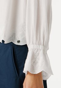 White long sleeve blouse with embroidered scalloped cuffs and hem, worn with dark blue pants with side pockets.