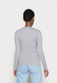 Osoba z krótkimi kręconymi włosami, ubrana w dopasowaną jasnoszaro długą koszulę i jasnoniebieskie dżinsy, odwrócona tyłem na jednolitym tle.