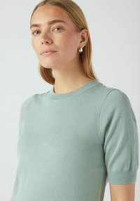 Svijetlozeleni kratkolisni pleteni džemper s okruglim izrezom, koji se odlikuje glatkom teksturom i suptilnim rebrastim detaljima na rukavima.