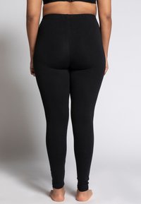 Leggings di cotone neri con una vita elasticizzata, forma aderente e texture liscia, mostrati da dietro con i piedi nudi.