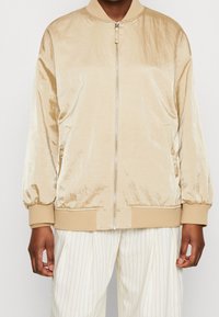 Chaqueta bomber de color beige claro con una textura suave y brillante, puños y dobladillo acanalados, y cremallera frontal. Los bolsillos laterales están integrados en el diseño.