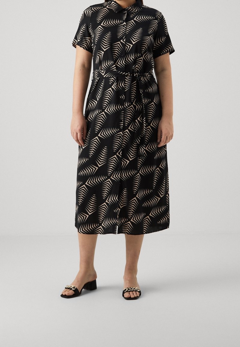 Femme portant une robe chemise midi noire avec des motifs géométriques de feuilles beige, accompagnée de sandales à talons blocs noires à bouts ouverts ornées d'une chaîne dorée.