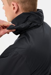 Veste noire avec un col haut, en tissu texturé, dotée d'une fermeture éclair et d'un col ajustable. Le design est épuré et fonctionnel.