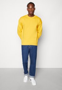 Gul överdimensionerad sweatshirt med rund hals, i kombination med blå denimjeans och vita sneakers. Enkelt, avslappnat outfit med luftig passform.