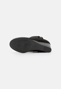 Chaussure en daim noir avec un embout arrondi, semelle en caoutchouc et un petit talon. Présente une boucle sur le côté pour un maintien sécurisé. Texture lisse tout au long.