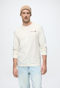 Camiseta de manga larga beige claro hecha de algodón, con un pequeño logo colorido en el pecho, combinada con jeans azul claro.