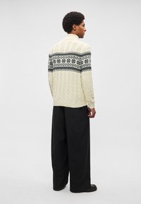 Suéter de malha com tranças creme com design preto estampado no peitoral, usado com calças pretas de perna larga. O suéter apresenta punhos e barra canelados.