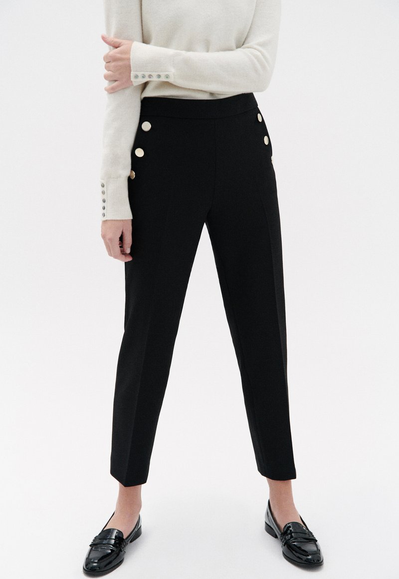 Zwarte op maat gemaakte broek met een slim fit, voorzien van gouden knoopaccenten op de tailleband en een gladde textuur. Gestyleerd met een lichte trui.