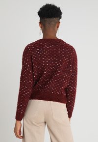 Pullover bordeaux fluffy con macchie multicolori, scollatura rotonda e orlo a coste. Indossato con pantaloni color crema. La texture sembra morbida.