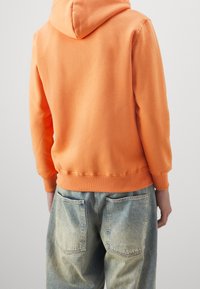 Felpa arancione con cappuccio e una texture morbida, con polsini e orlo a costine, abbinata a jeans in denim blu sbiaditi con due tasche posteriori.