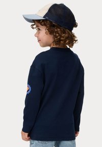 Marinblå långärmad skjorta med rund halsringning och en dekorativ patch på vänster arm, tillsammans med en blå och krämfärgad baseballkeps.