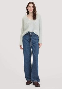 Hellgrüner, gerippter Strickpullover mit V-Ausschnitt. Dunkelblaue Jeans mit weiter Beinform und Bindegürtel. Braune Schuhe runden das Outfit ab.