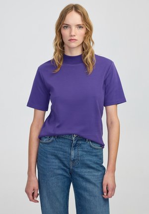 Paarse t-shirt met korte mouwen en hoge hals, gemaakt van een gladde stof, gecombineerd met lichtblauwe high-waisted jeans, op een neutrale achtergrond.