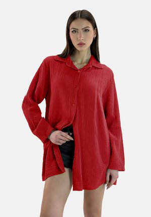 Chemise rouge texturée à manches longues avec un col, fermeture à boutons sur le devant et fente sur les côtés, accompagnée de shorts noirs.