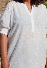 Persona con capelli ricci che indossa una blusa bianca a maniche corte con texture, scollatura a V e polsini smock, in piedi all'aperto alla luce del sole.