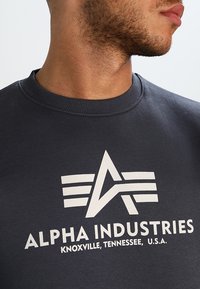Felpa grigia scura in morbido tessuto, decorata con un grande logo stampato bianco che comprende il testo "ALPHA INDUSTRIES" e elementi di design.
